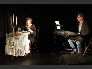 Ewa Dałkowska siedzi przy stoliku z mikrofonem w ręku. Z prawej strony Rafał Gajewski przy instrumencie klawiszowym.