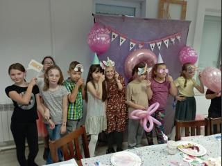 10-osobowa grupa gości pozuje do zdjęcia z zabawnymi gadżetami. W tle różowa ścianka i balony urodzinowe.