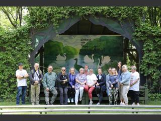 Kadra Polany Kultury pozuje do wspólnego zdjęcia przy scenie w Leśniczowce Pranie. Z tyłu na scenie namalowane zielone gesi. W tle drzewa.
