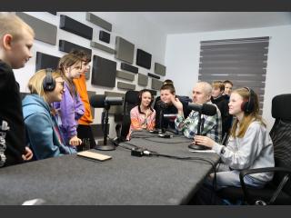10 dzieci, młodzieży, oraz mężczyzna w Polskim Radiu Orzysz. Trzy dziewczyny siedzą na krzesłach, mają na głowie słuchawki, przed nimi mikrofony. Obok nich instruktor - Pan Krzysztof Roszko, dookoła nich reszta dzieci.