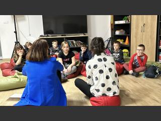 Na podłodze na pufach siedzą dwie kobiety - Pani Monika Samul oraz Pani Agnieszka Racis, przed nimi siedzą dzieci.