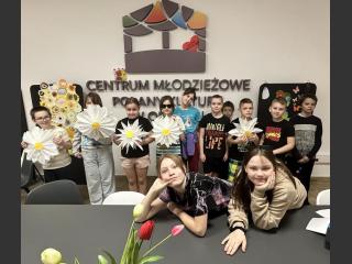 Dzieci pozują do wspólnego zdjęcia z dużymi kwiatami. Za nimi logo Centrum Młodzieżowego.