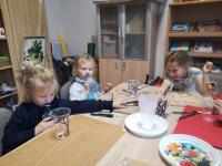 Przy stole trzy dziewczynki. Dwie dziewczynki z lewej strony piją wodę z butelek. Na stole pomalowane przez nie pastikowe kubki. 