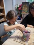 Dziewczynka siedzi przy stole. W rękach trzyma pomalowany przez siebie plastikowy kubek. 