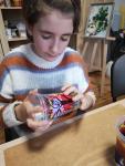 Dziewczynka siedzi przy stole, trzyma w rękach pomalowane przez siebie plastikowy kubek. Za nią na sztaludze obraz.