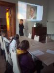 Dzieci w sali w Pałacu Kultury w Orzyszu podczas zajęć z języka angielskiego. Dzieci siedzą przy stole, przed nim kartki. Przed nimi Pani Dyrektor Polany Kultury- Joanna Kamieniecka.