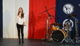 Dziewczyna na scenie, trzyma w ręku mikrofon, śpiewa. Z prawej strony na scenie statyw mikrofonowy, perkusja. W tle flaga Polski.