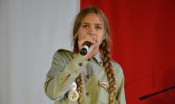 Dziewczynka trzyma mikrofin, śpiewa. W tle lafa Polski. Przegląd piosenki partiotycznej i żołnierskiej.