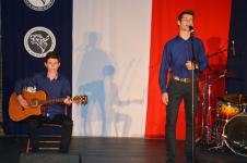 Dwóch mężczyzn na scenie. Po lewej mężczyzna siedzi na krześle i gra na gitarze, po prawej meżcyzna stoi przed statywem mikrofonowym, śpiewa. Z tyłu perkusja. W tle flaga Polski, logo z tygrysem.