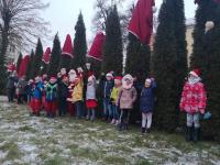 Dzieci pozujące do zdjęcia z Panią Basią przy drzewach przustrojonych w czapki św. Mikołaja.