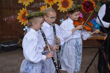 Trzy dziewczynki na scenie/ Dwie cziewczynki z lewej strony graja na klarnetach. Dziewczynka po prawej stronie gra na skrzypcach. W tle słoneczniki. XIII Orzyskie Spotkanie Folklorystyczne