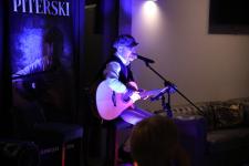 Piotr Huegel Piterski z gitarą, przed nim statyw mikrofonowy, na scenie w restauracji Toscana w sercu Mazur.