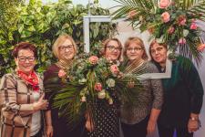 Z lewej strony stoja dwie kobiety, obok nich trzy kobiety pobujące do zdjęcia w ramie ozdobionej kwiatami. Za nimi na ścianie rośliny. W restauracji Toscana w sercu Mazur.
