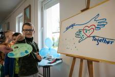 Chłopiec pokazuje do zdjęcia kciuka w niebieskiej farbie. Z prawej strony obraz z dłońmi i napisem "STRONGER TOGETHER"
