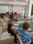Przy stołach dzieci uczą się się kodowania za pomocą puzzli ukladają drogę dla robota. Z tyłu przy jednym ze stołów Pani mgr Małgorzata Cwalina. Z tyłu na ścianie wyświetlany obrazek.