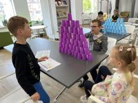 Trzy grupy dzieci przy stołach układają uwieżę z kolorowych, plastikowych kubków. W Sali konferencyjnej w Bibliotece Polany Kultury.