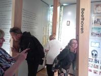 Pracownicy Polany Kultury w Muzeum K.I. Gałczyńskiego. Stoją z głowami przy drewnianych belkach, w środku z głośnikami i słuchają muzyki klasynczej. Z lewej strony kobieta robi zdjęcia. Na ścianach napisy, zdjęcia.