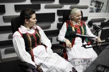 Dwie kobiety w strojach ludowych w studiu Polskiego Radia Orzysz, przed nimi statywy mikrofonowe.