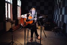W studiu nagrań kobieta siedzi na krzesle, gra na gitarze, na głowie ma sluchawki, przed nią dwa statywy mikrofonowe 