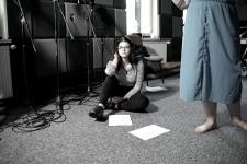 W studiu nagrań kobieta siedzi na podłodze, obok widać nogi drugiej osoby. Z lewej strony przy ścianie stoja statywy mikrofonowe