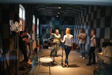 Sześć osób w studiu nagrań. Stoją w kole, przed nimi statywy mikrofonowe. w trakcie nagrań do płyty "Leć głosie po rosie".