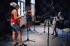 Cztery kobiety w studiu nagrań. Z prawej strony kobieta gra na gitarze. Z tyłu perkusja 