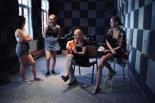 Cztery kobiety w studiu nagrań. Dwie siedzą na krzesłach, druga kobieta z prawej gra na gitarze