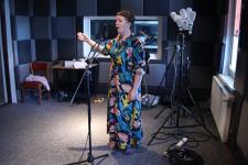 Kobieta w studiu nagrań, śpiewa, przed nią statyw mikrofonowy, za nią lapa. 