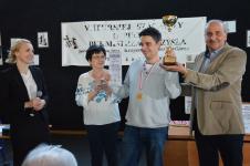 Mężczyzna trzyma w ręku puchar i dyplom, na szyji ma medal za zajęcie pierwszego miejsca w turnieju szachowym. Z prawej strony Wicestarosta Piski, z lewej strony Pani Dyrektor Polany Kultury.