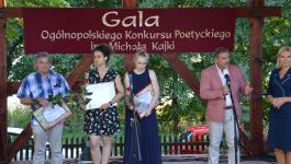 Pięć osób na scenie. Przy sooby po lewej trzymają ramki z dyplomami i róże. Po prawej stronie stoi Burmistrz Orzysza Zbigniew Włodkowski, przed nim statyw mikrofonowy.