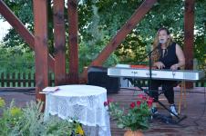 Kobieta na scenie gra na keyboardzie i śpiewa. Przed nią stolik nakryty obrusem, kwiaty w wazonie i koszu.