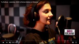 Zrzut ekranu z filmu na YouTube. W studiu nagrań, dziewczyna śpiewa, przed nią mikrofon.